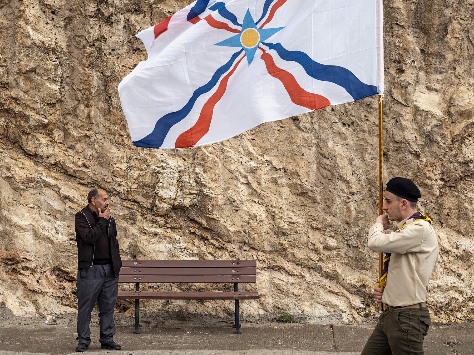 In Between — a man and a young flag bearer face each other beside a bench, an Assyrian flag overhead