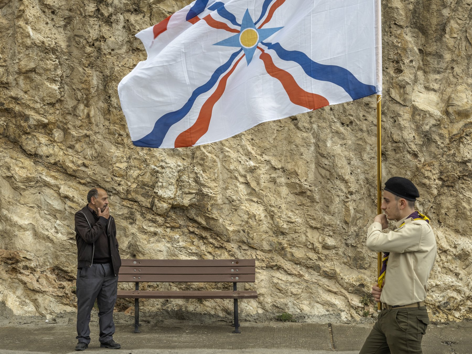 A man and a young flag bearer face each other beside a bench, an Assyrian flag overhead, Duhok, Iraq
