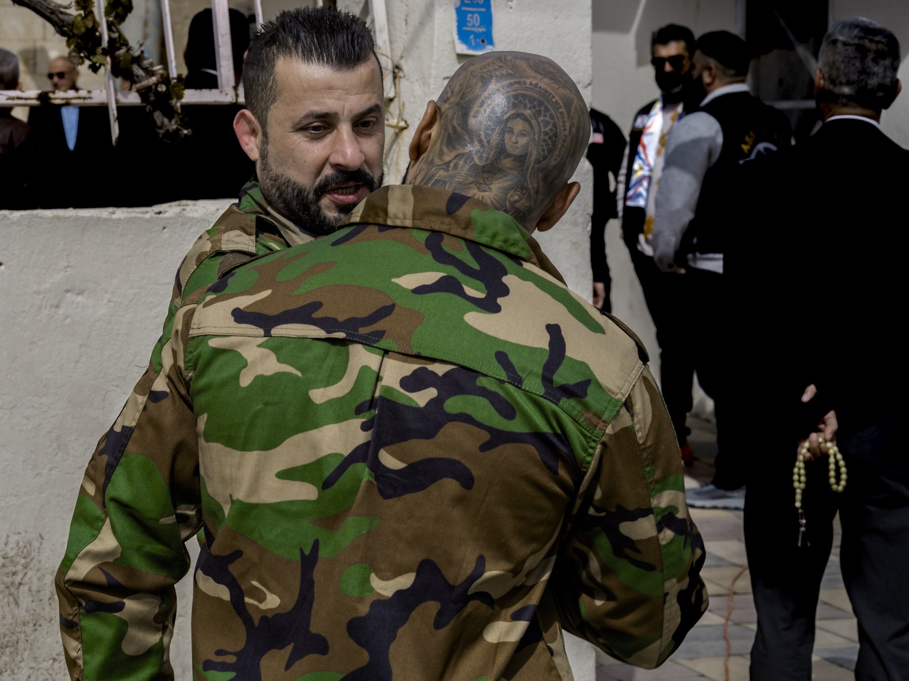 A man in camouflage embraces another man whose shaved head bears an elaborate religious tattoo