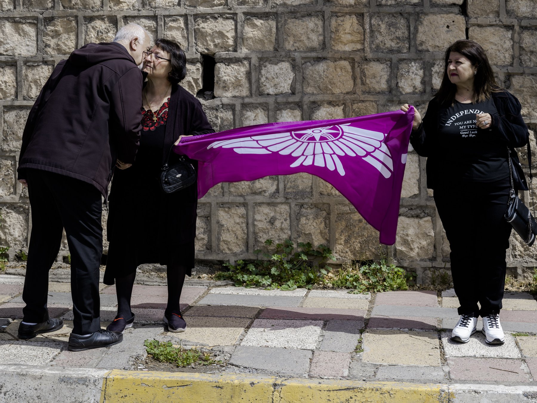 Three people exchange a greeting beside a purple flag with a winged emblem, against a stone wall