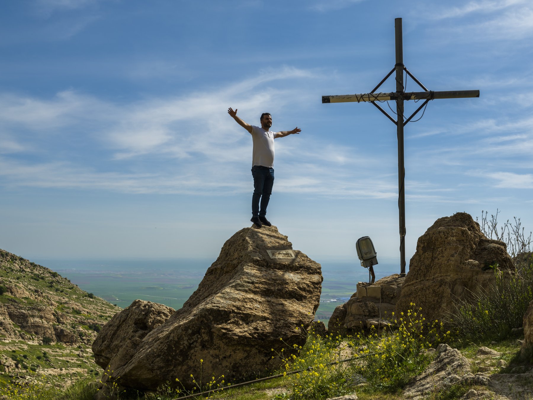 A man stands with arms outstretched atop a large boulder beside a tall cross, overlooking a valley