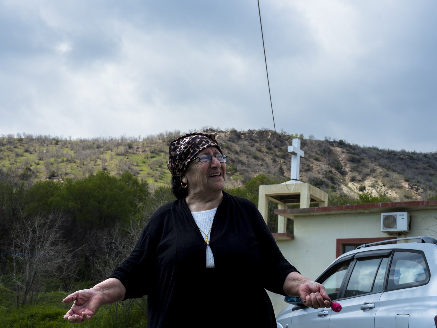 An older woman in a headscarf stands smiling outdoors, a hilltop cross and church behind her