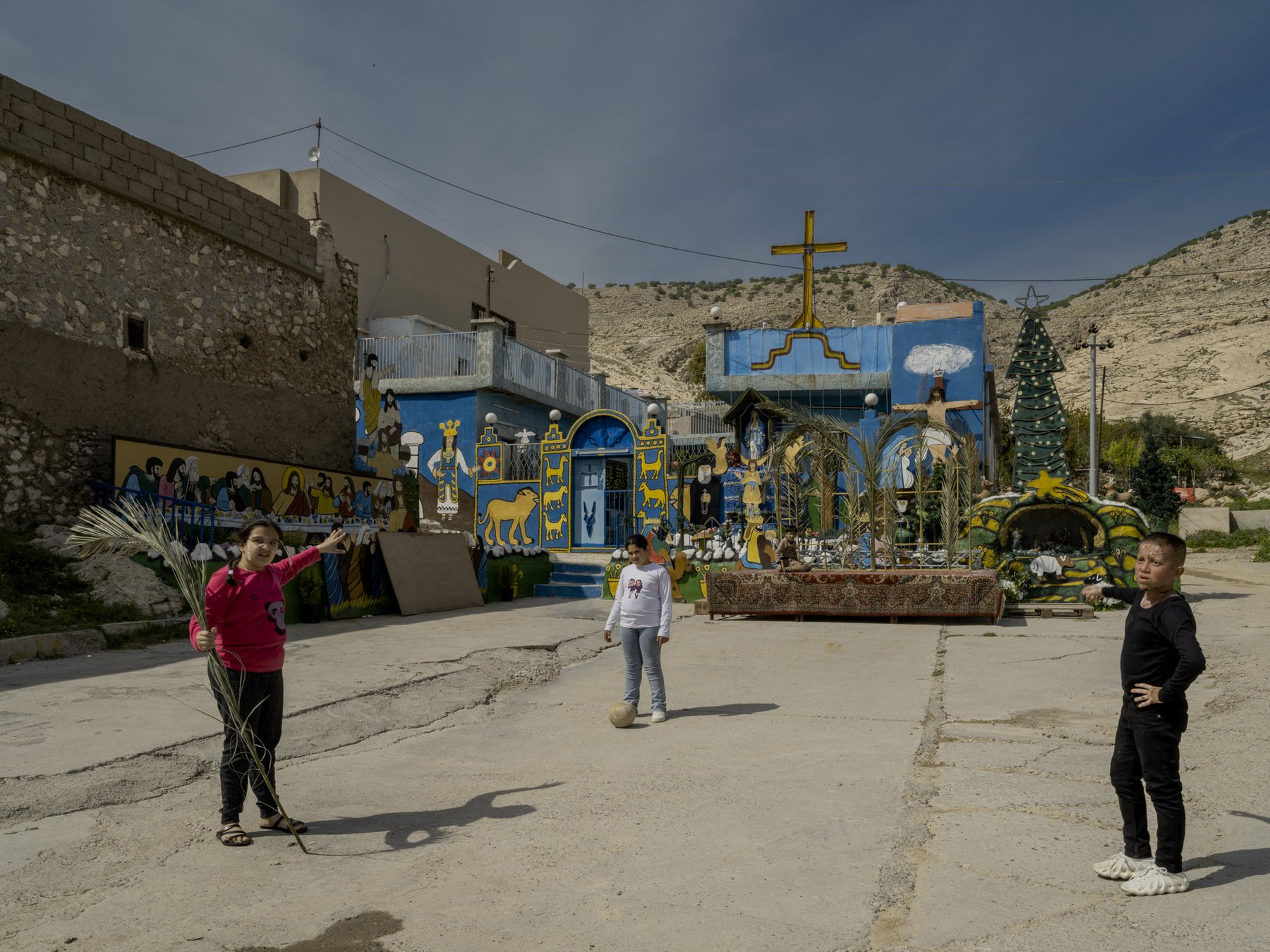 Children play with a ball and palm fronds in front of a brightly painted religious mural