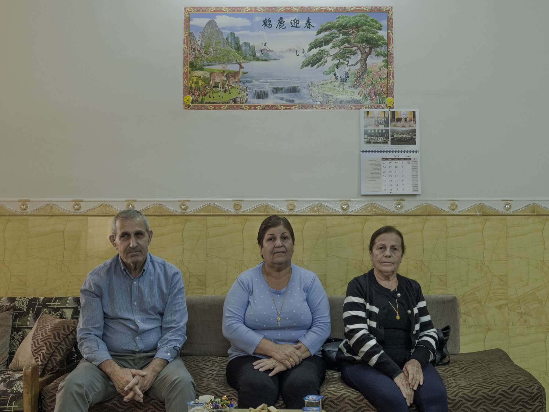 Three people sit in a row on a sofa beneath a framed landscape painting and a calendar