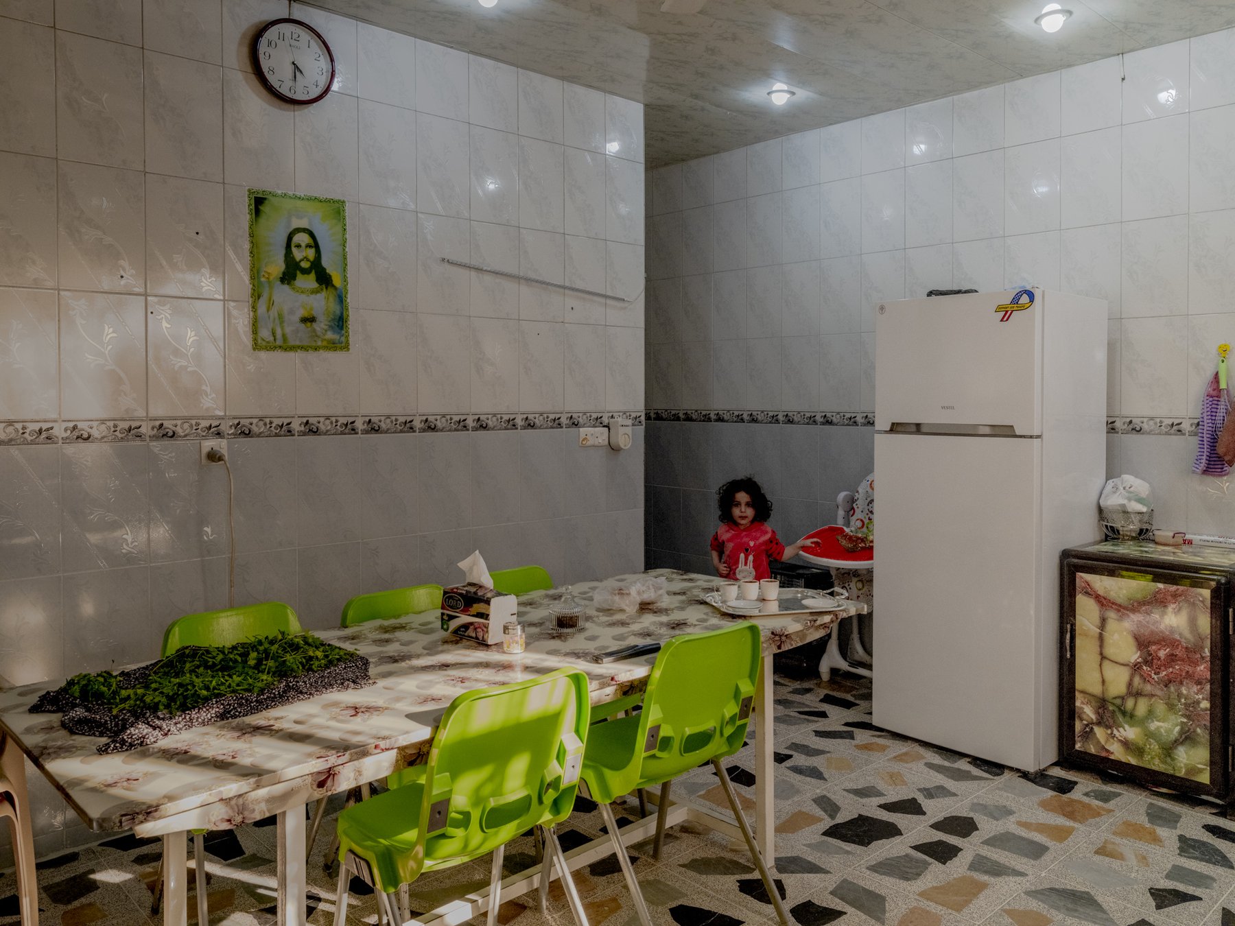 A young child stands in a tiled kitchen beside a refrigerator and a table set for tea