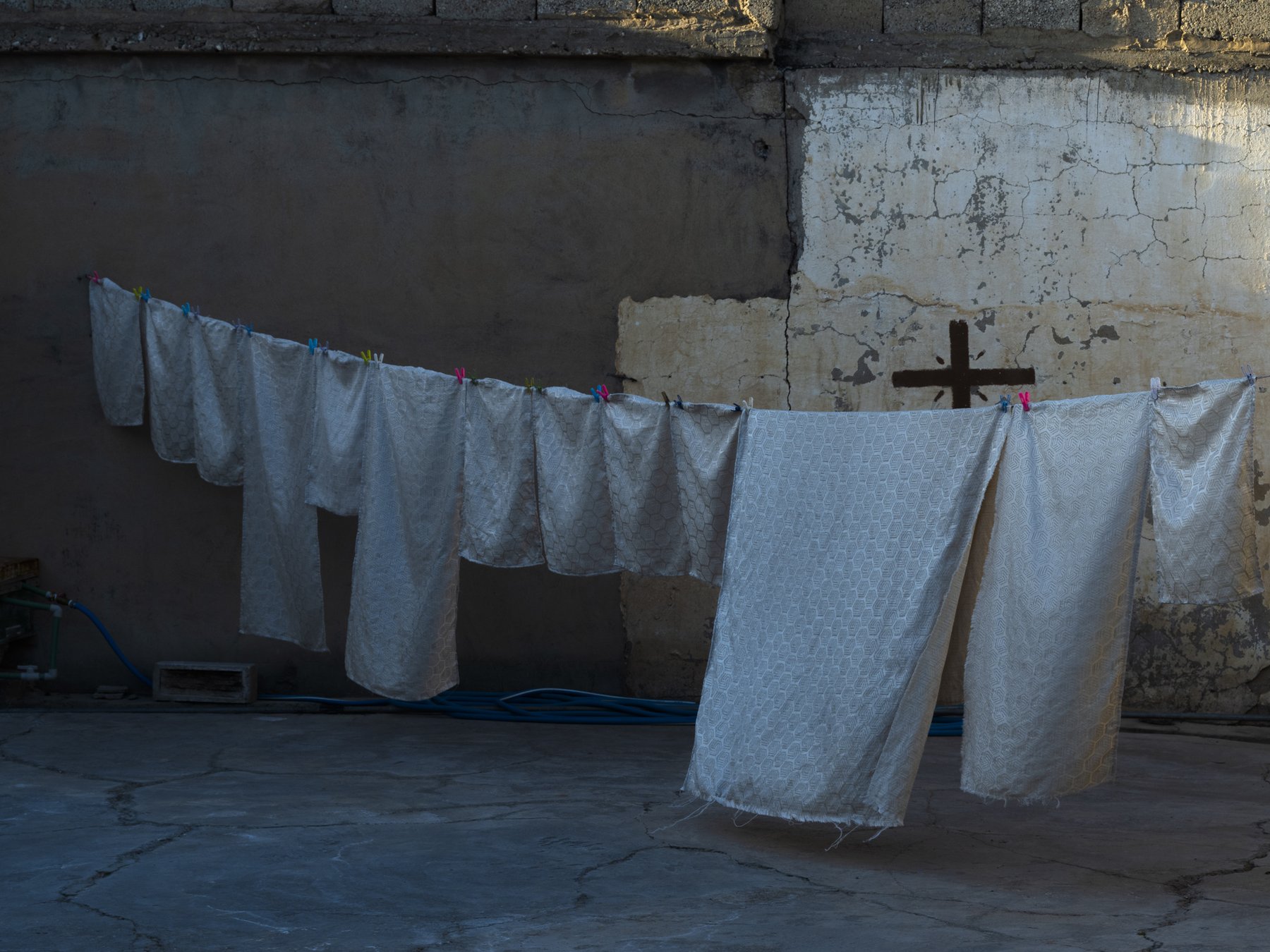 White patterned cloths hang to dry on a line against a weathered wall marked with a painted cross
