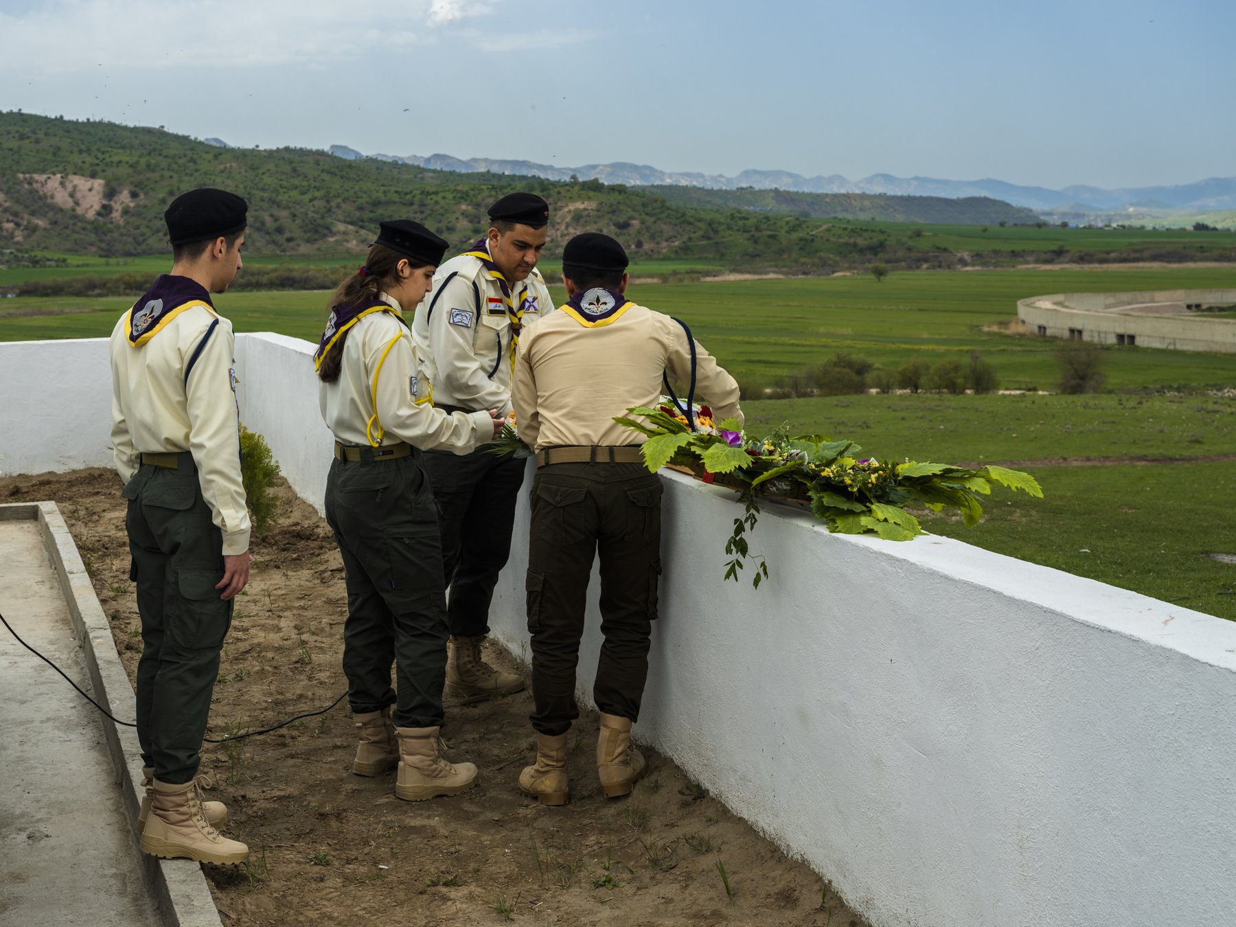 Four young people in scout uniforms arrange flowers along a low wall overlooking a green valley