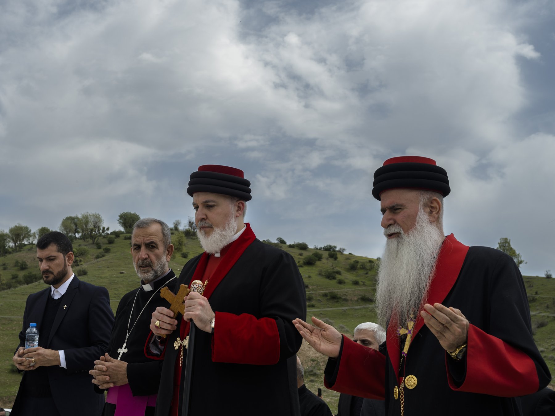 Two clergy members in red-and-black vestments pray outdoors beneath a cloudy sky