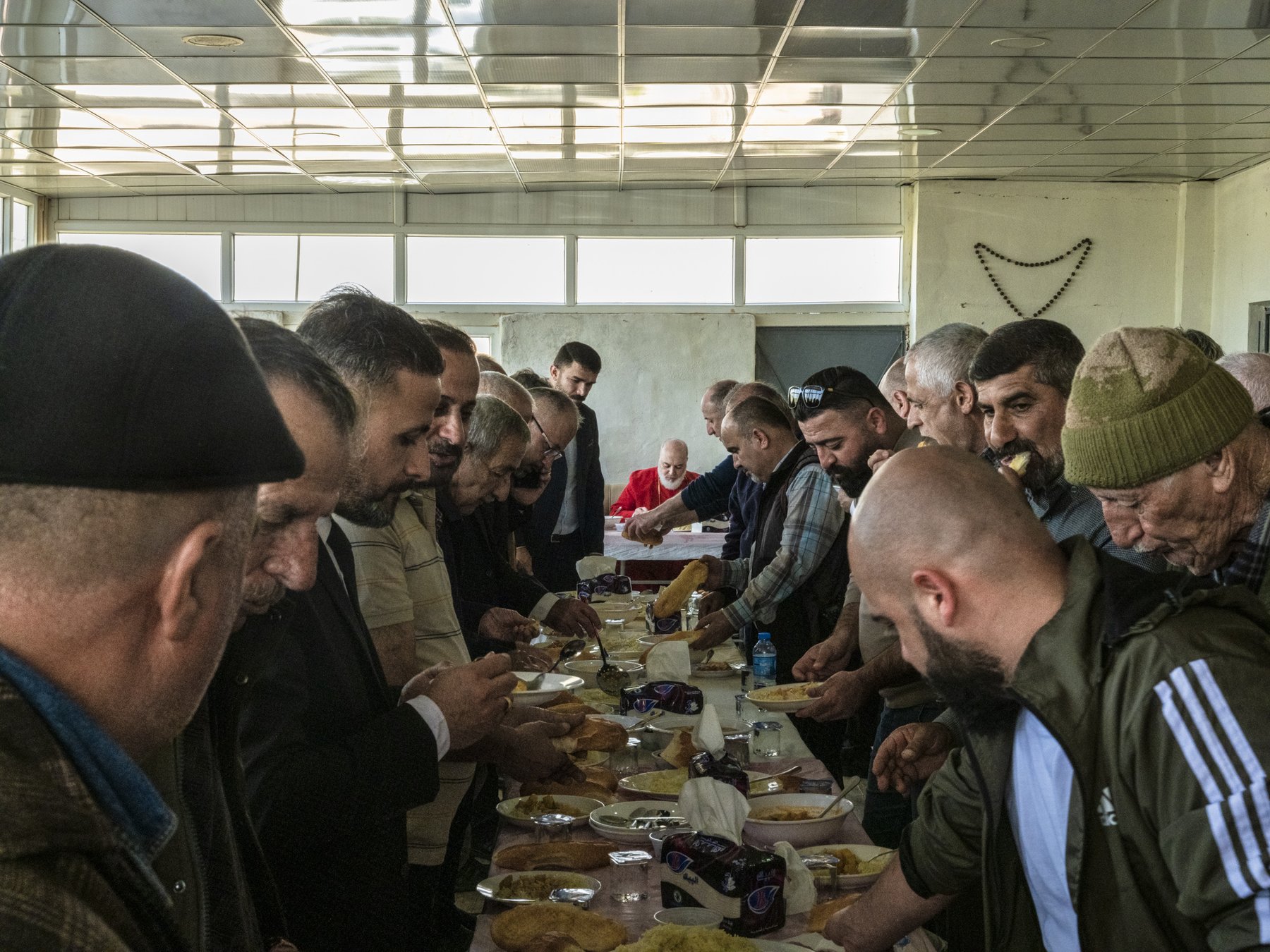 A long line of men serve themselves food from a communal table inside a hall