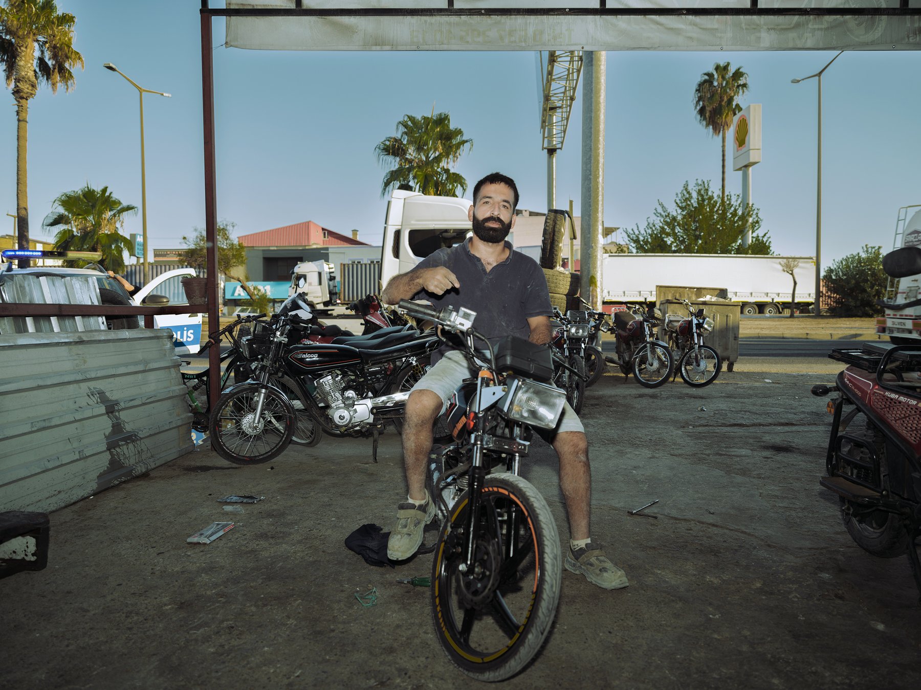 A bearded man sits on a motorcycle under a roadside canopy, palm trees and trucks behind him