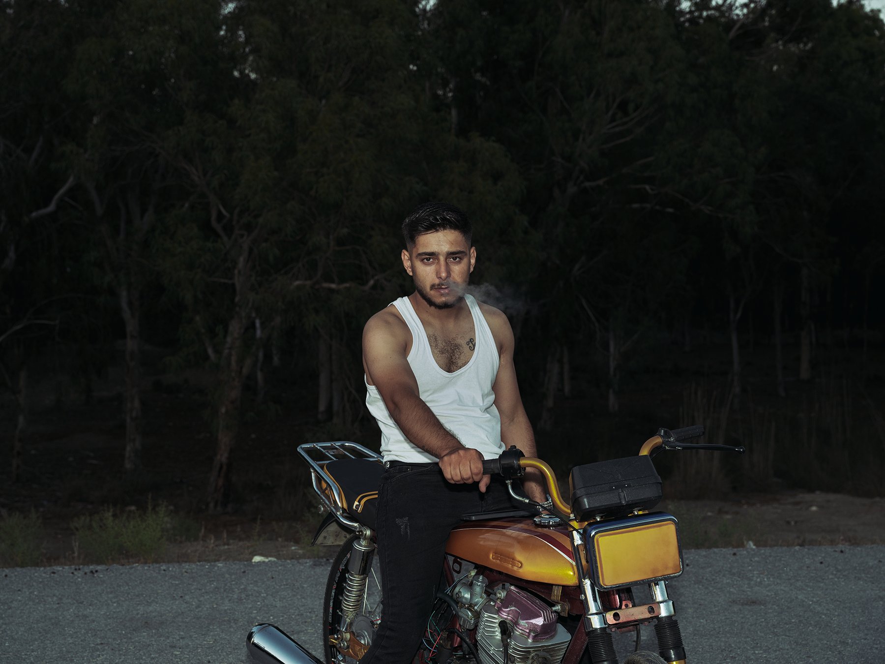 A young man in a white tank top sits on a customized motorcycle at dusk, exhaling smoke