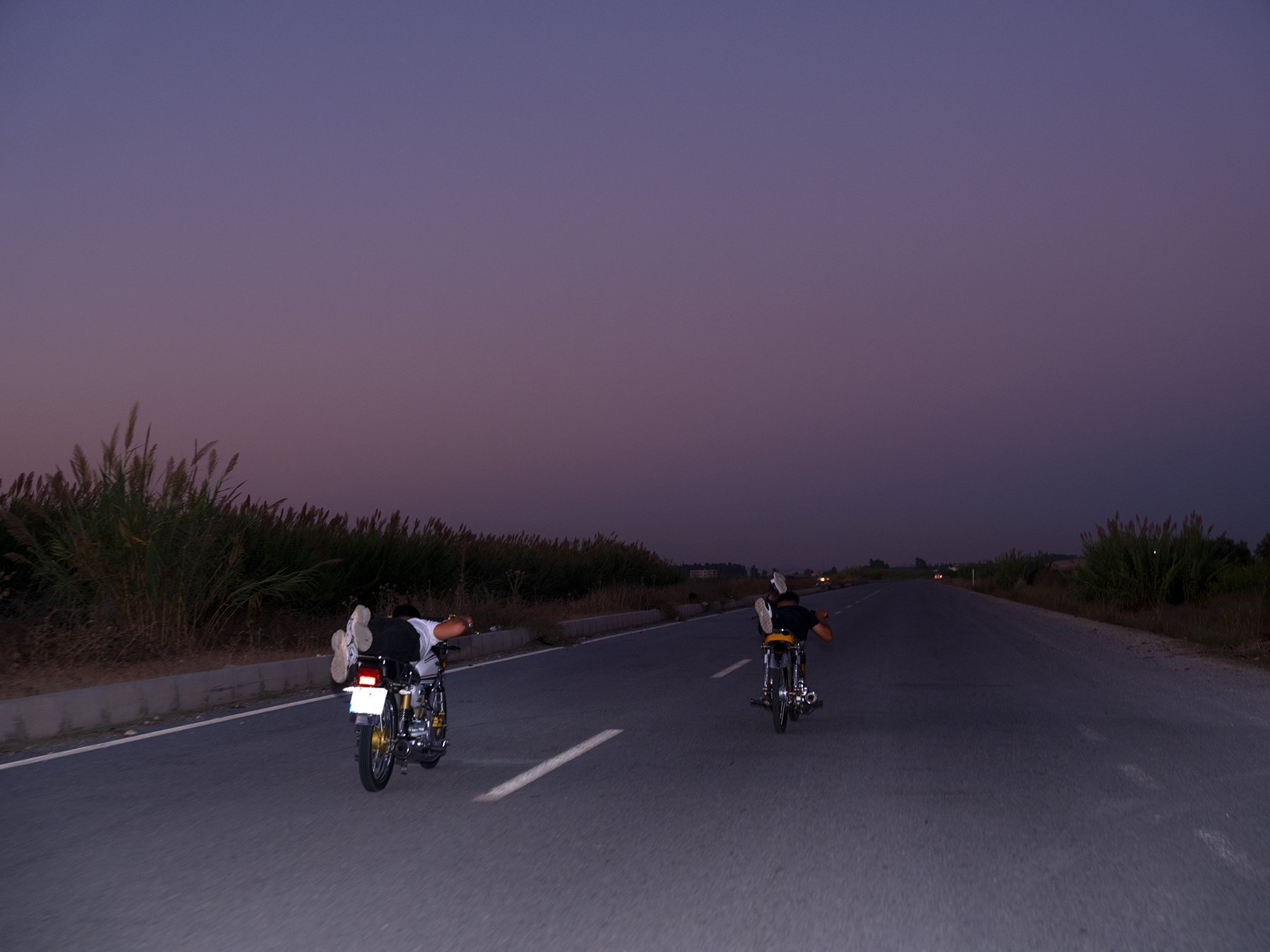 Two riders lie flat across their motorcycles, speeding down a road at dusk