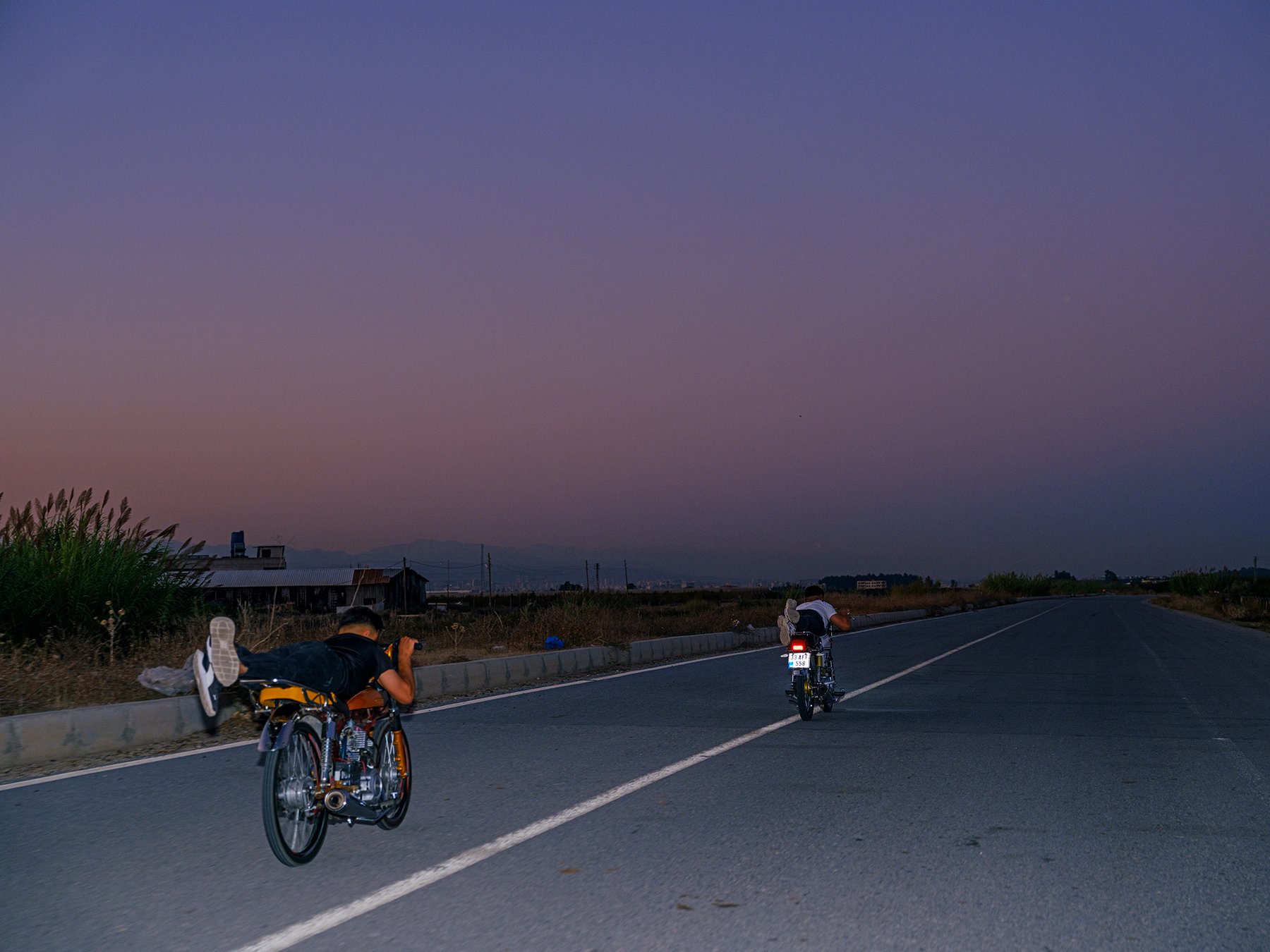 Two riders lie prone on their motorcycles, riding away down a road at dusk