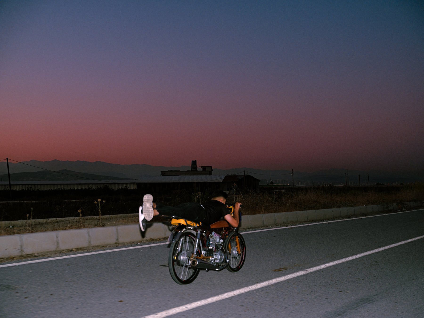 A rider lies flat across a customized motorcycle mid-stunt on a road at dusk