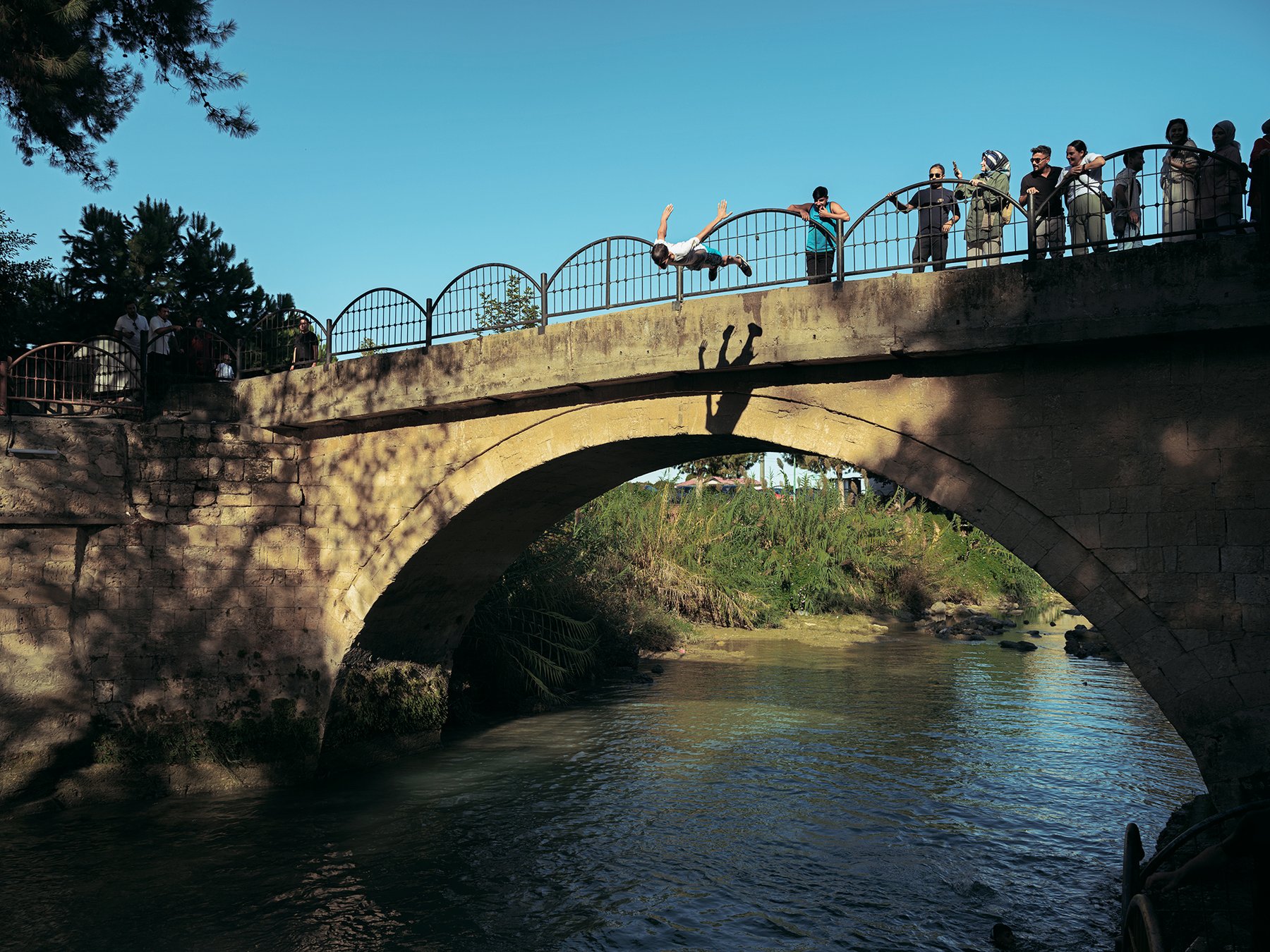 A young person flips through the air off a stone bridge into a river, onlookers watching
