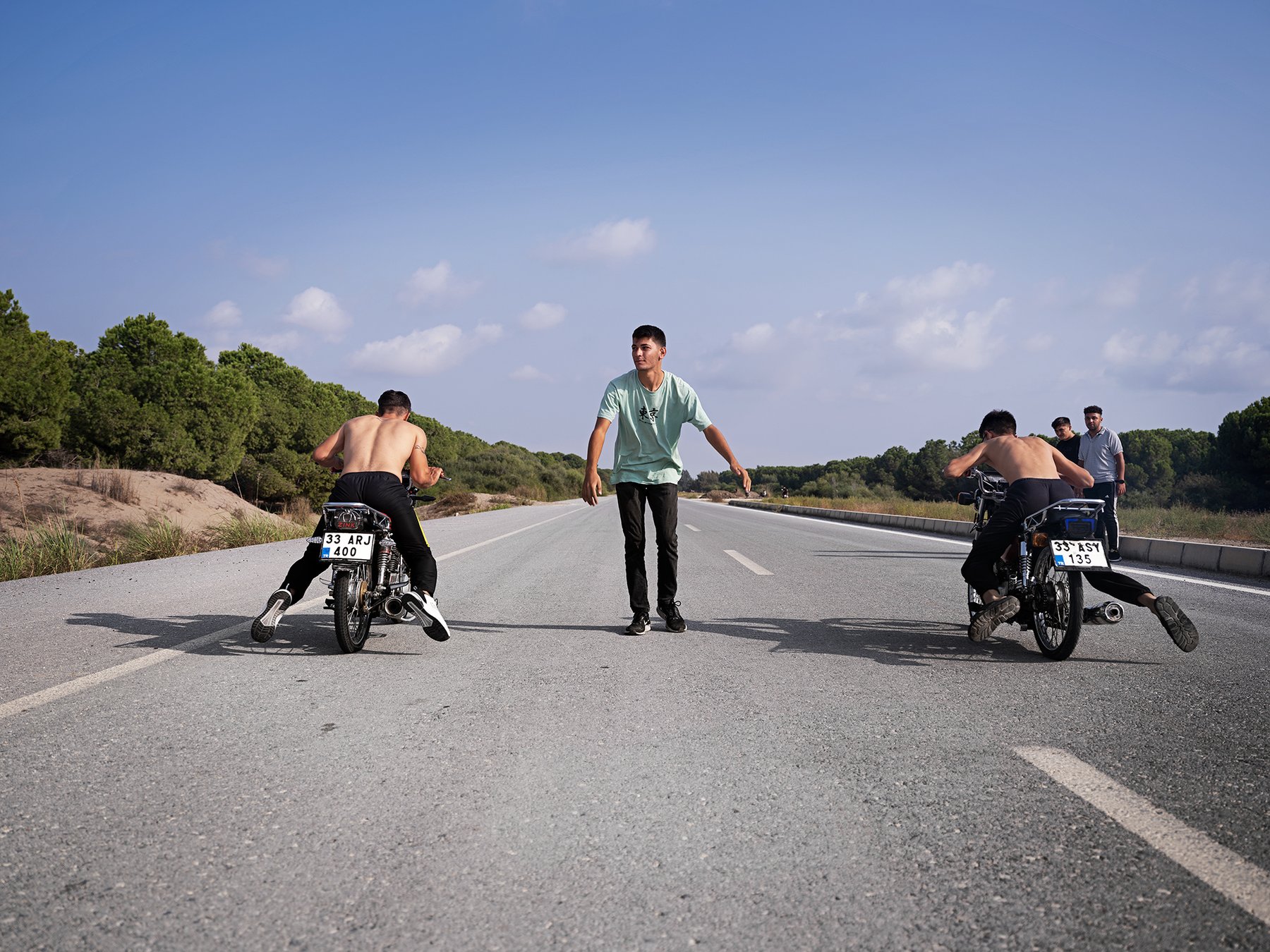 Two riders lie flat across their motorcycles, riding away down a long straight road