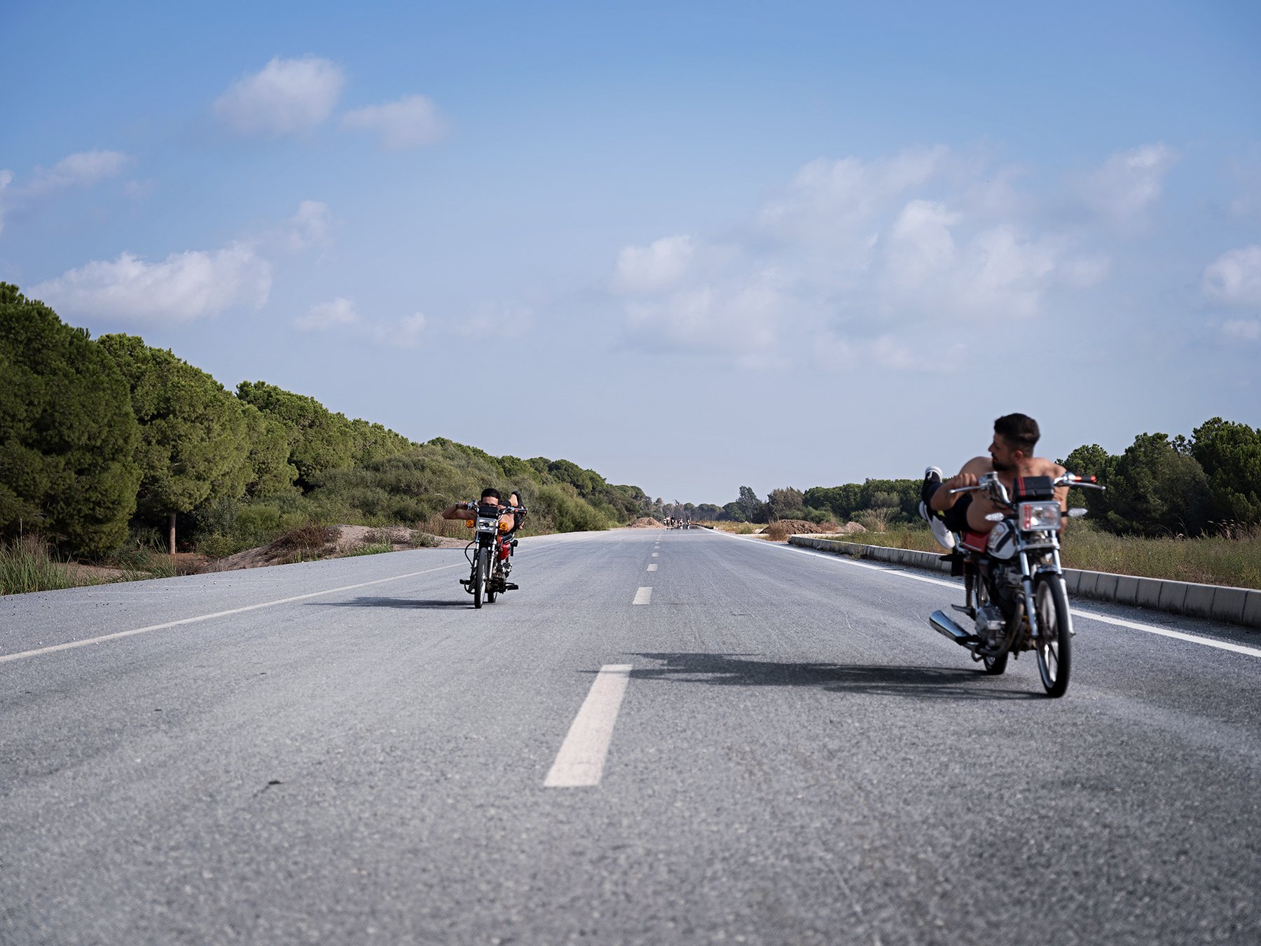 A man in a white tank top stands with his customized motorcycle on an empty road