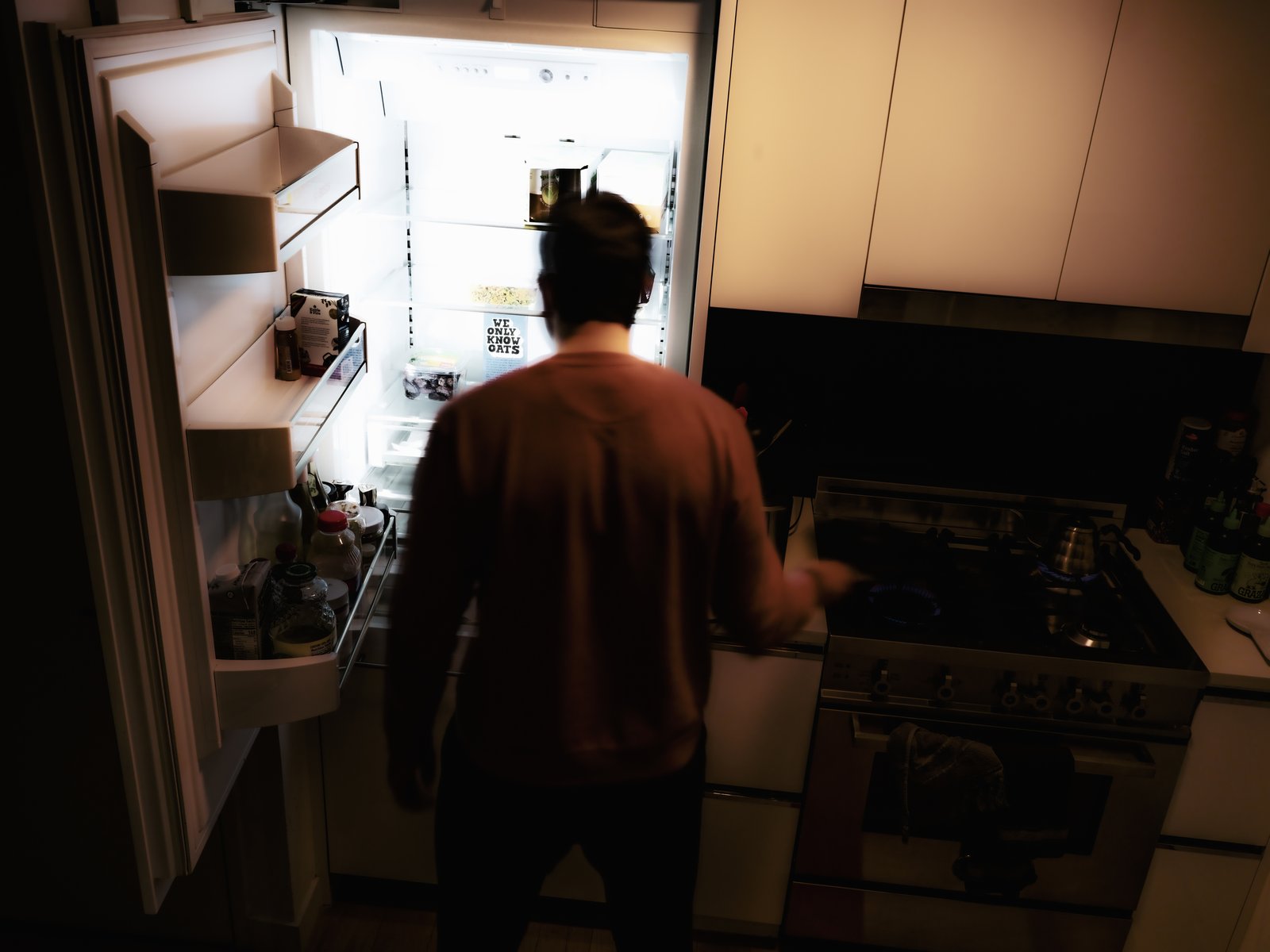 A man stands before an open, brightly lit refrigerator at night, his back to camera