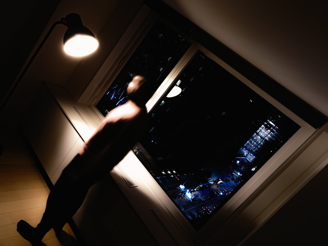 A blurred figure stands in a dim room beside a lit lamp, a night cityscape through the window