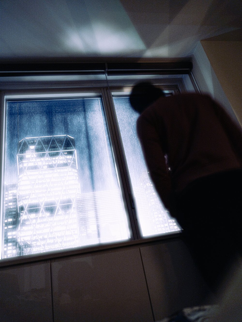A blurred silhouette beside a rain-streaked window overlooking an illuminated angular skyscraper