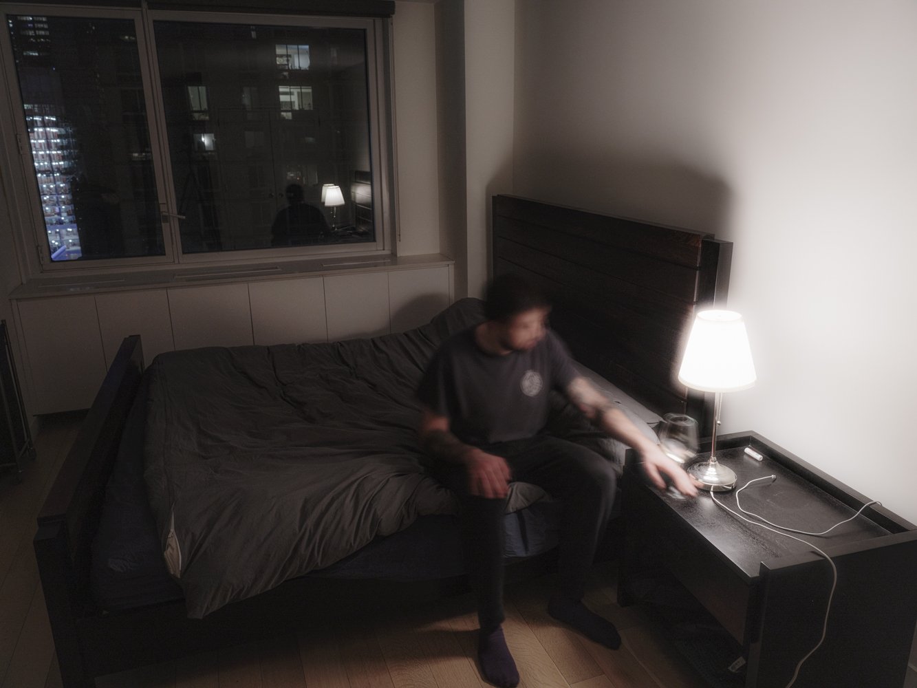 A man sits on the edge of a bed reaching toward a bedside lamp, his reflection caught in the window beside a glowing city skyline
