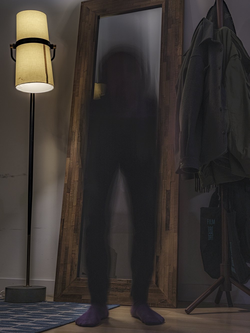 A blurred, ghostly silhouette stands before a full-length mirror beside a lit floor lamp and a coat rack