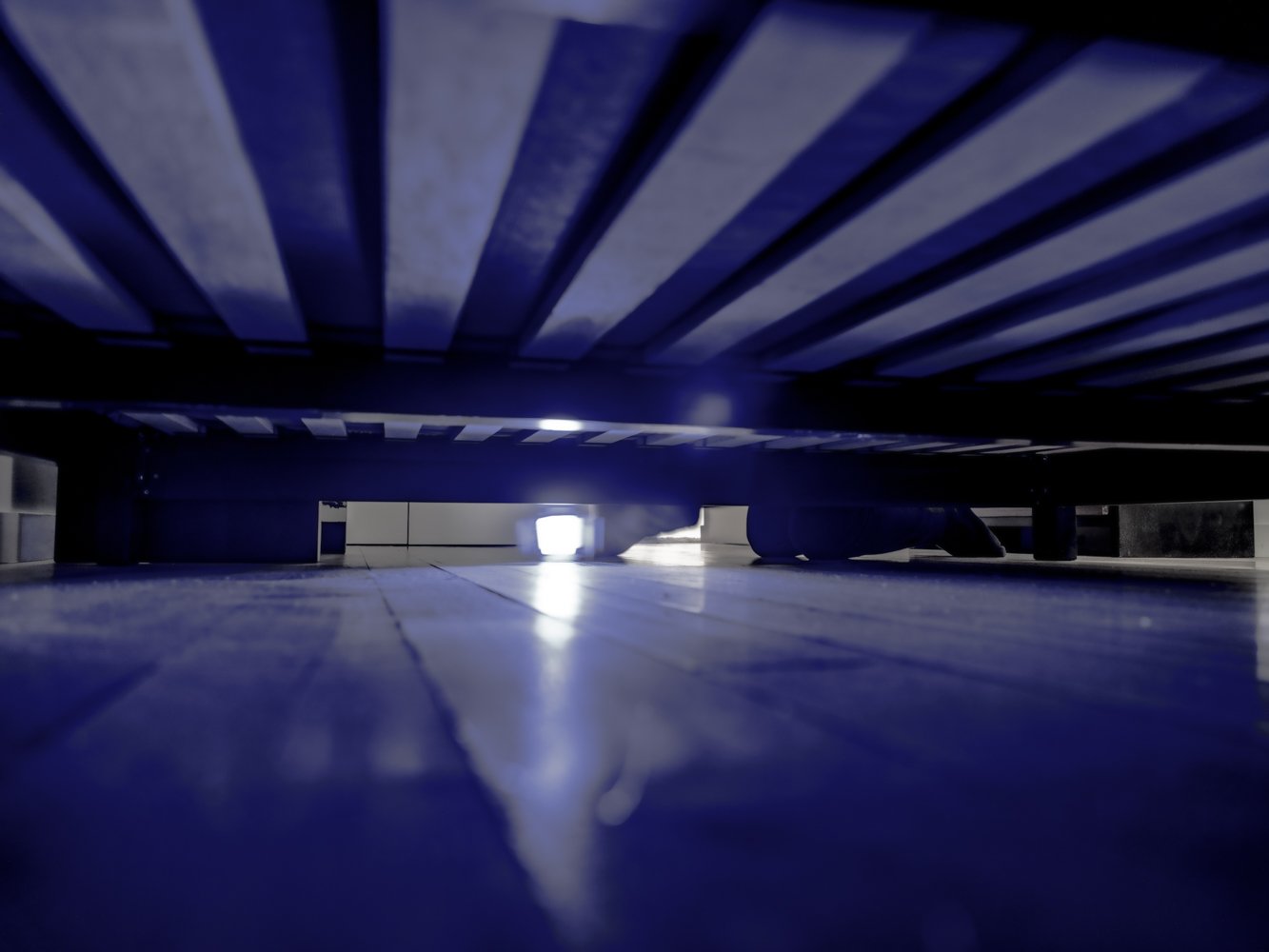 A low, blue-toned view beneath a bed frame toward a glowing light source