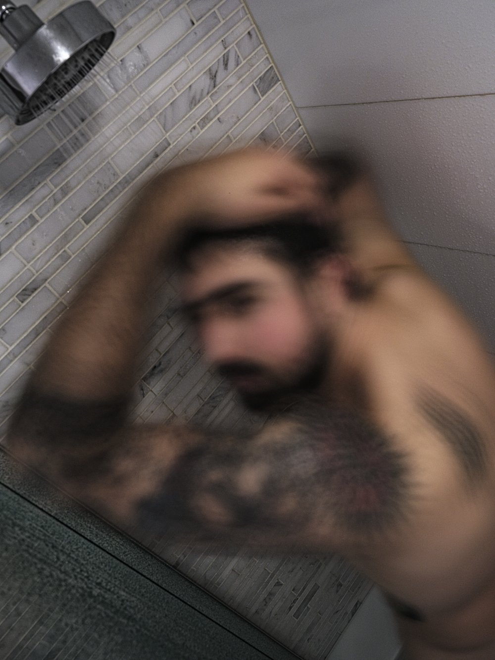 A blurred close-up of a tattooed man washing his hair under a shower head