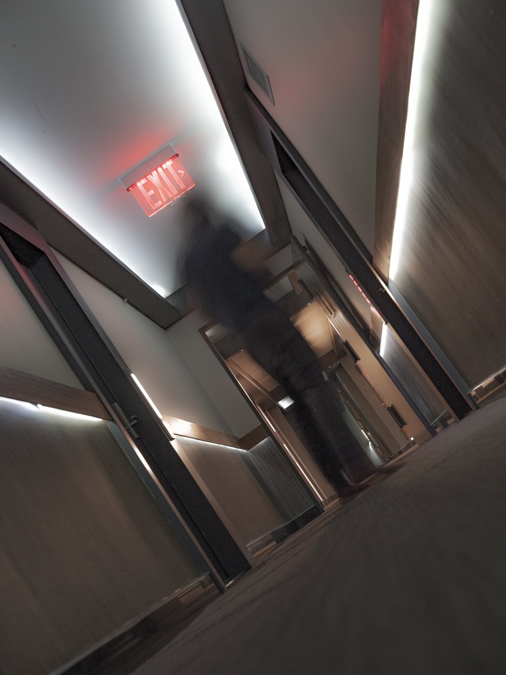 A blurred figure walks down a hotel-like corridor beneath a glowing red EXIT sign