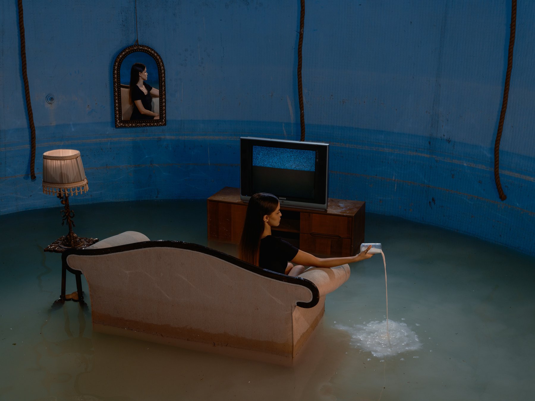 A woman sits on a flooded, leaning sofa watching a static-filled TV in a surreal blue-tinted room