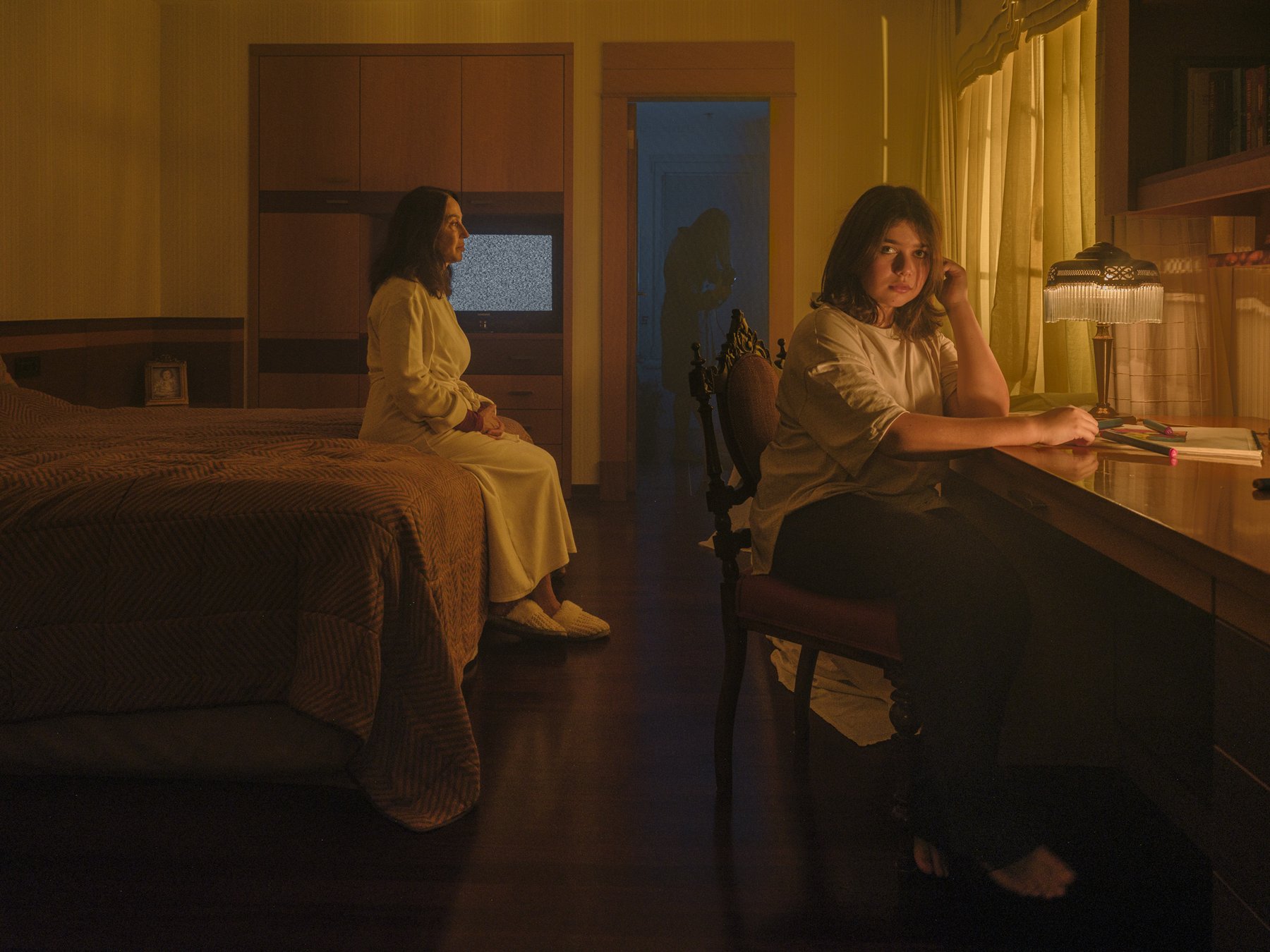 A woman sits on a bed before a static-filled TV while a younger woman sits separately at a desk in a warmly lit bedroom, a shadowed figure in the doorway