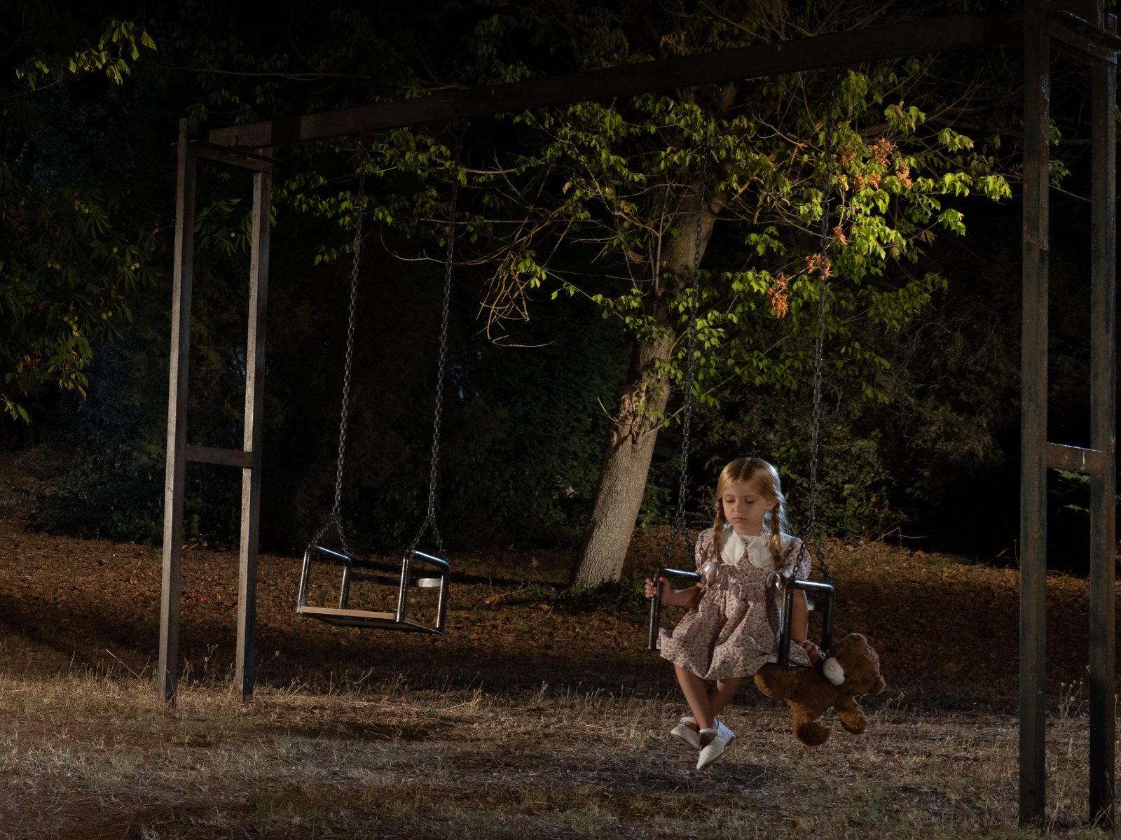 A young girl in a vintage dress sits alone on a playground swing at night, a teddy bear resting beside her