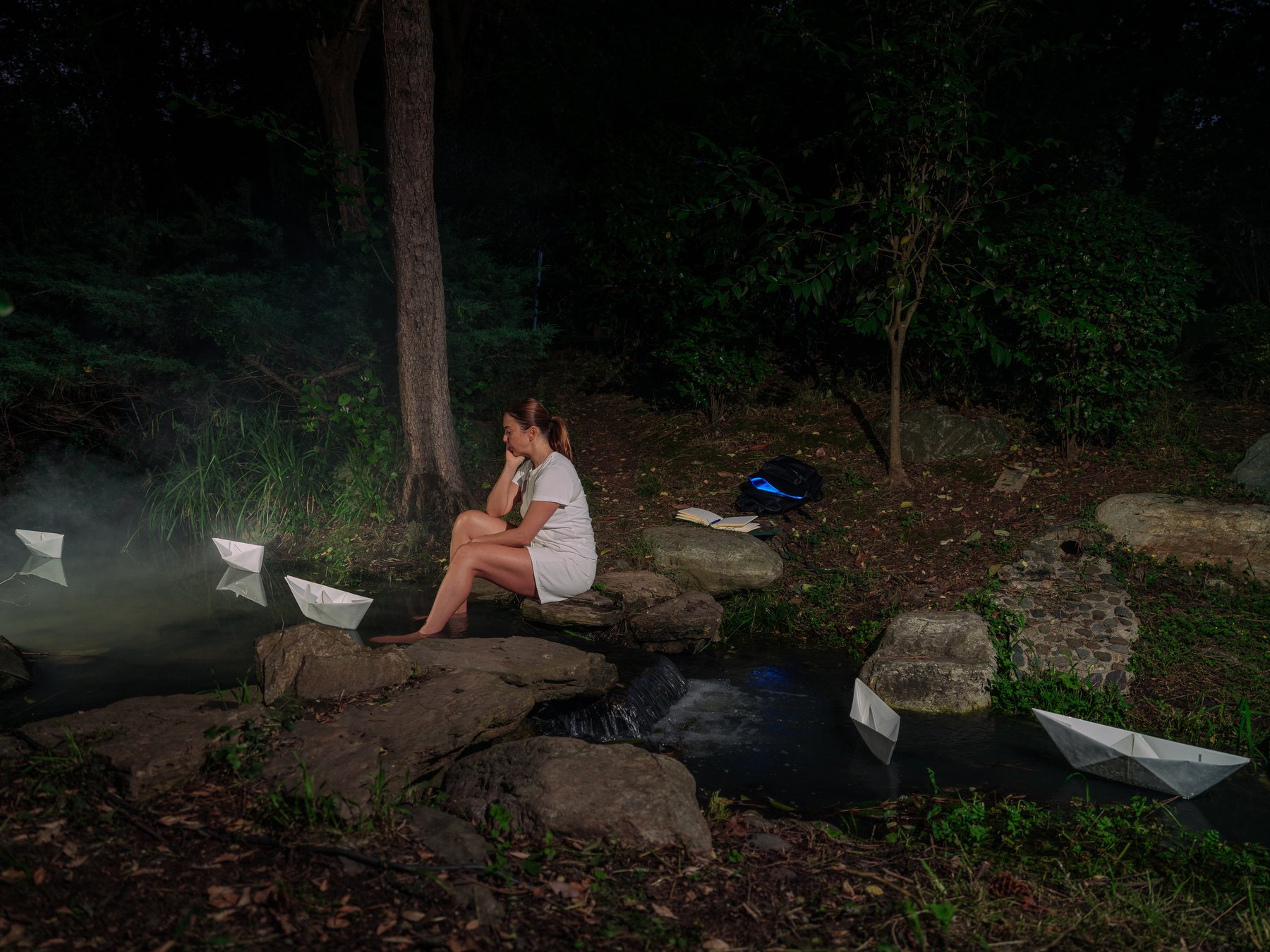 A woman sits pensively at the edge of a dark pond at night, paper boats floating on the water beside her
