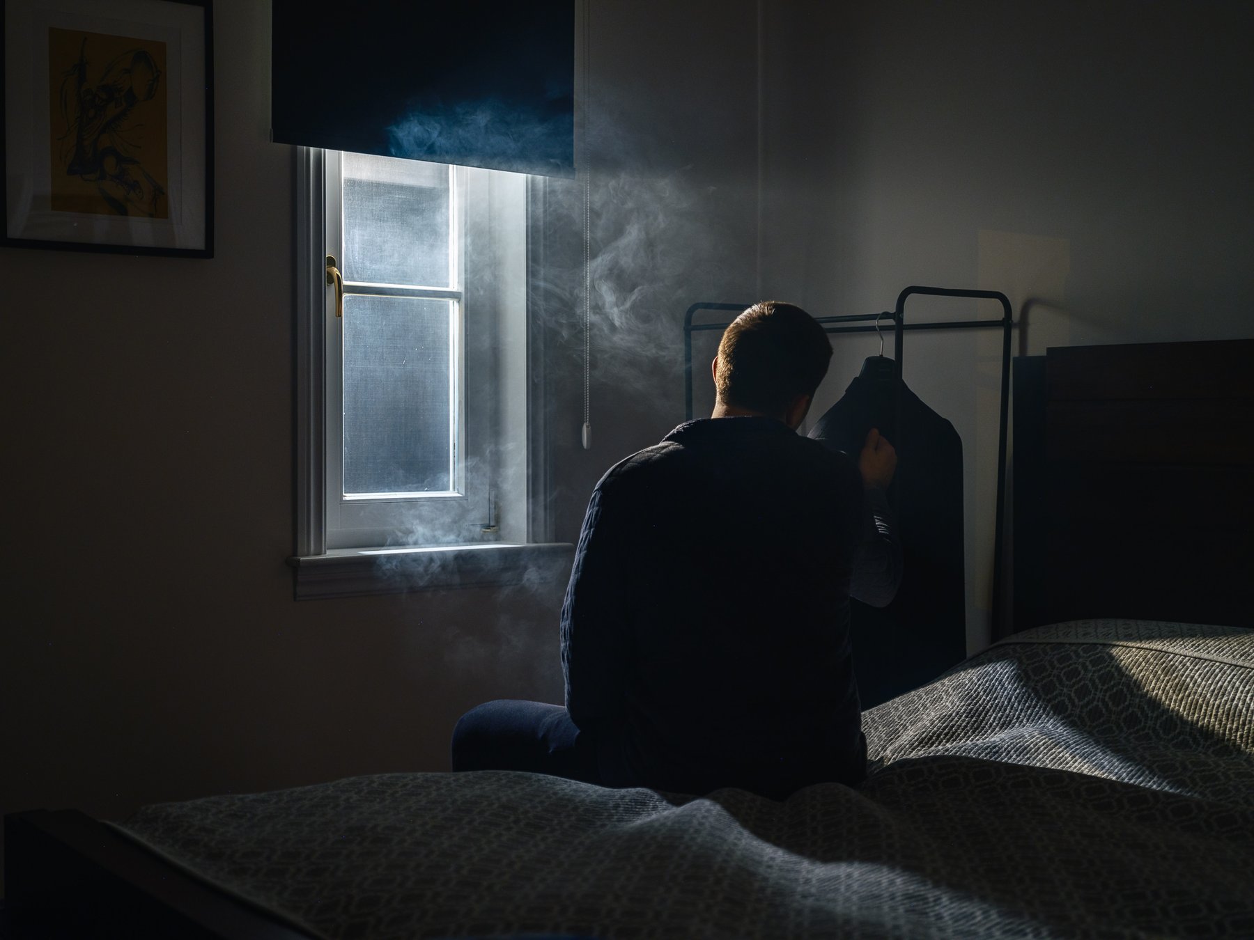A man's silhouette stands by a window filled with drifting smoke, reaching toward a dark garment hanging by the bed